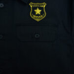 Close-up of a black security guard shirt with a yellow embroidered badge that reads SECURITY SERVICE and features a star.
