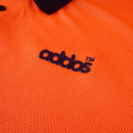 A close-up shot of an orange athletic polo shirt featuring a black embroidered knockoff logo that says addos with a TM symbol.
