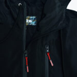 Close-up of a black hooded jacket featuring two zippers with bright red pull tabs and a brand label visible on the inside collar.