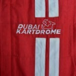 A close-up of a red racing suit featuring the white embroidered logo DUBAI KARTDROME with a racetrack outline and vertical white stripes.