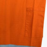 Close-up of bright orange workwear fabric showing a stitched rectangular pocket and a reflective blue strip at the bottom.