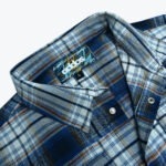 Close-up of a blue and brown plaid flannel shirt collar showing the Addos Workwear Collection label and metal snap buttons.