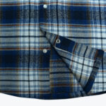 Close-up of a blue, white, and brown plaid flannel shirt showing the metal snap buttons and curved hem.