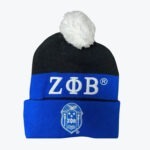 A black and blue knit beanie with a white pom-pom, featuring the white embroidered Greek letters ZΑB on the middle band and the Zeta Phi Beta shield crest on the folded cuff.
